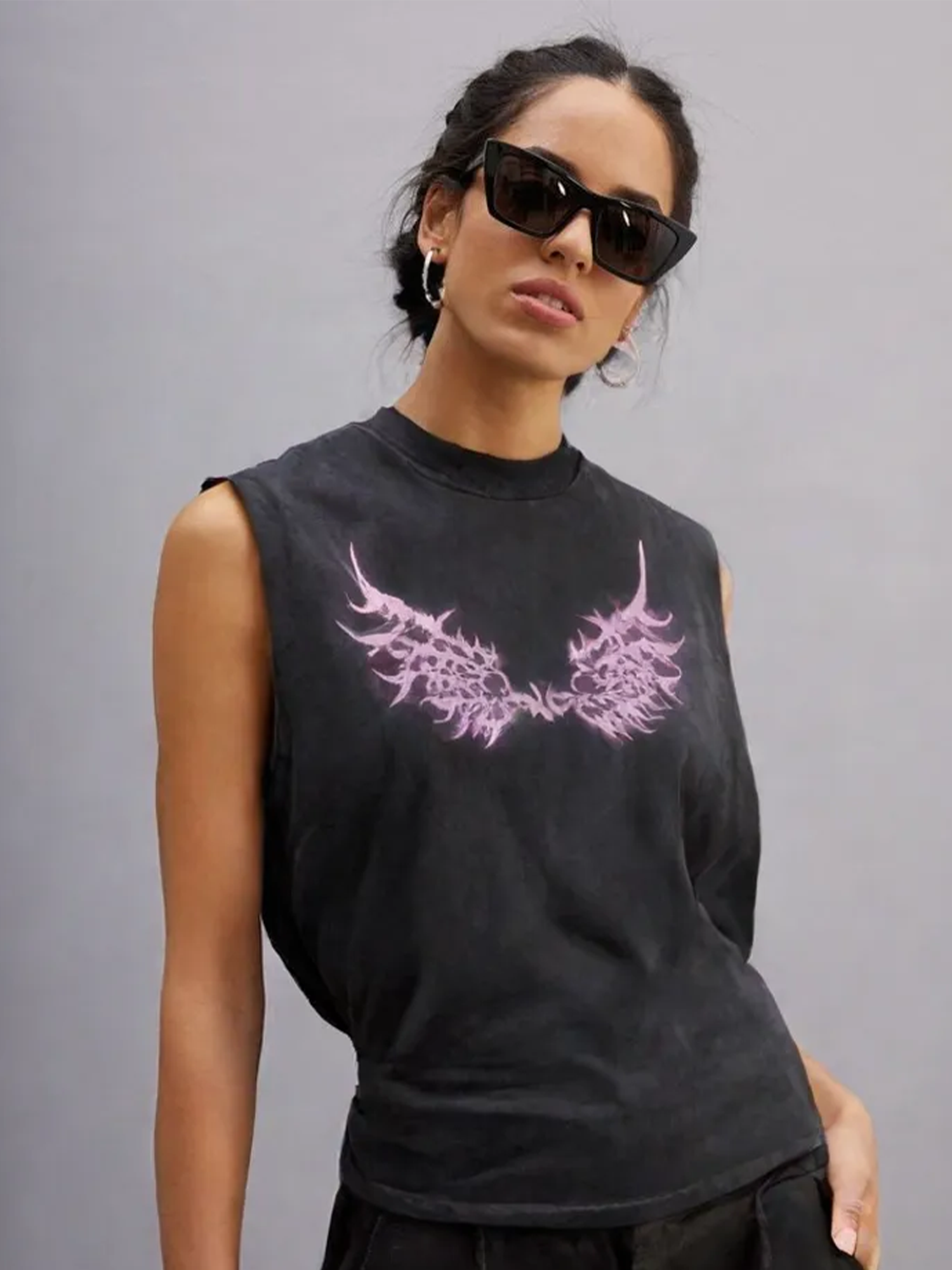 Woman wearing black sunglasses and a black sleeveless tank top with pink wing graphics printed on the chest, posed against a gray background