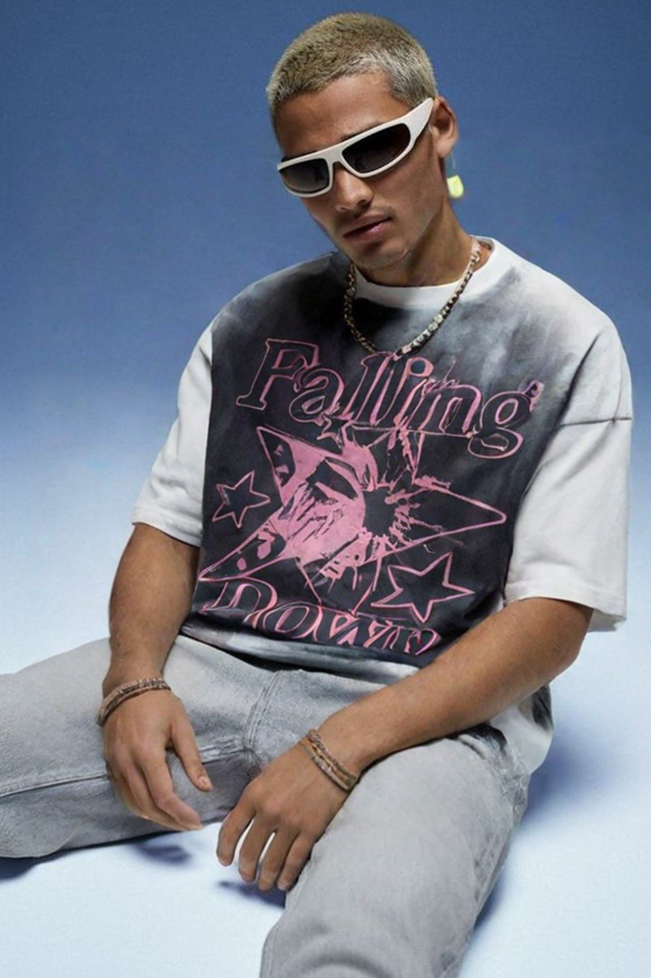 Person wearing white wraparound sunglasses and oversized t-shirt with 'Falling' text in pink lettering plus graphic design featuring stars and other decorative elements, paired with light gray jeans, sitting against gradient blue background