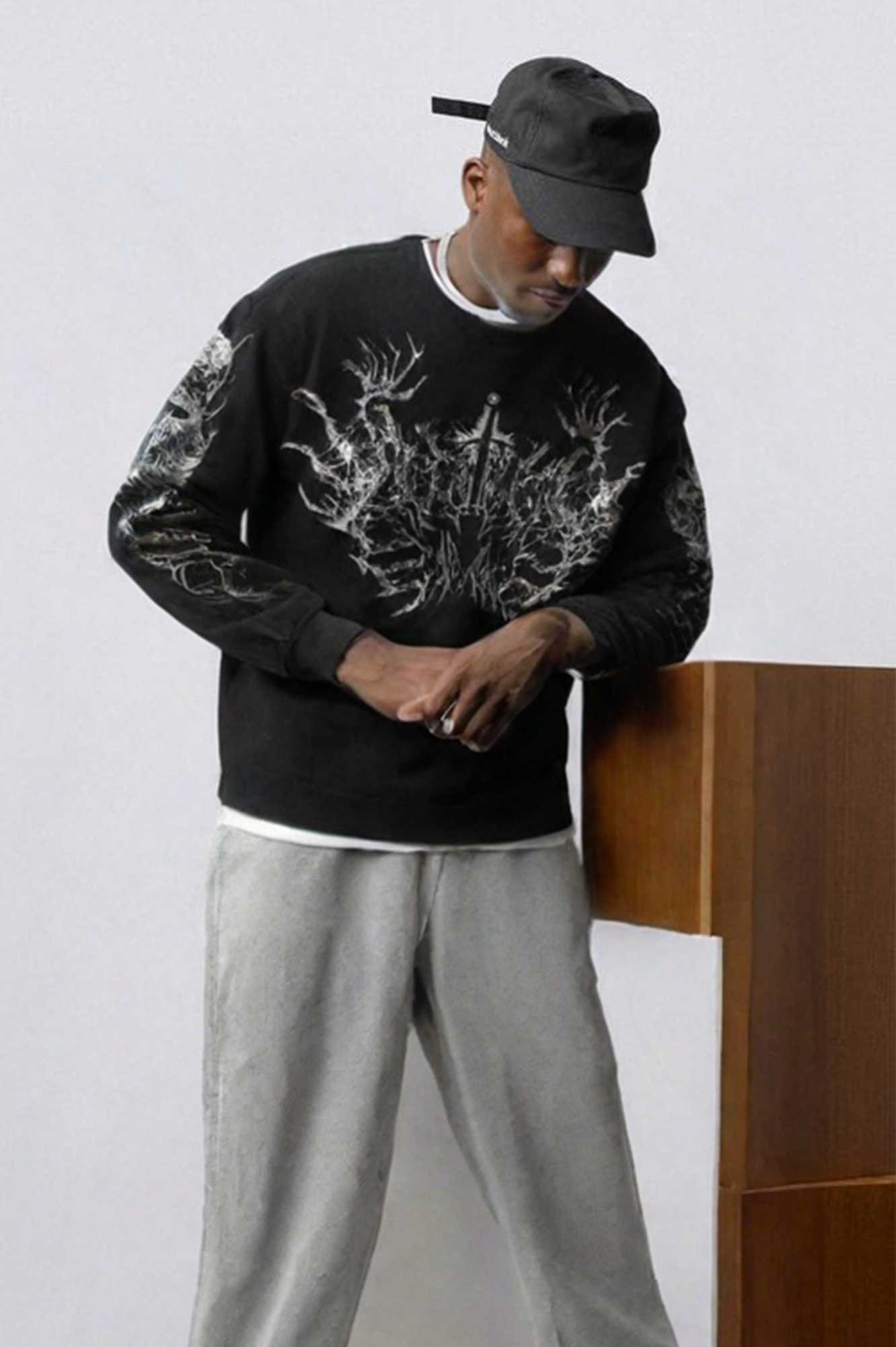 Person wearing black bucket hat, black sweatshirt with white botanical/tree branch graphic design on chest and sleeves, and gray sweatpants, standing indoors