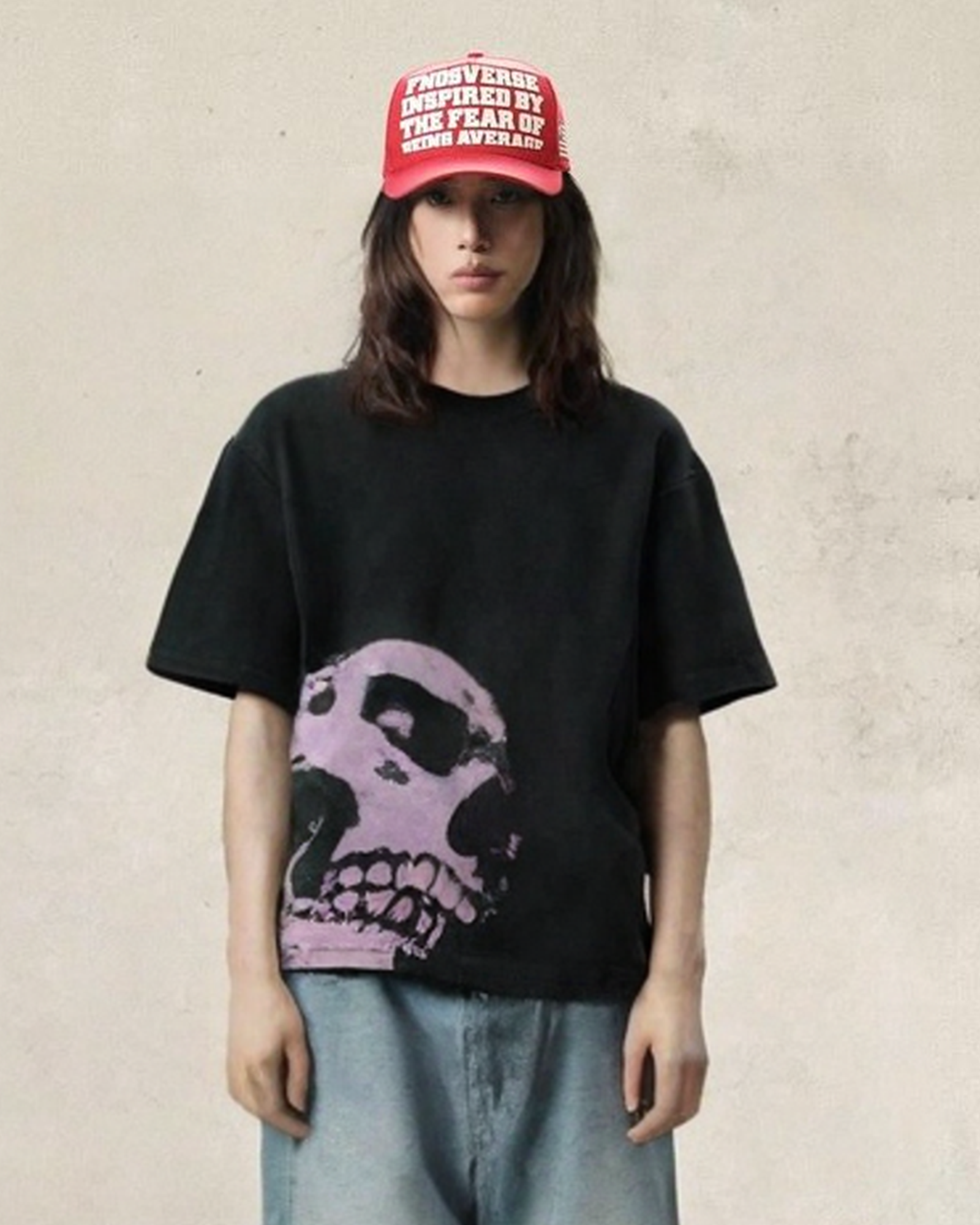 Person wearing red baseball cap with white text reading 'BOYS VERSUS GIRLS INSPIRED BY THE FEAR OF BEING AVERAGE', black oversized t-shirt with large pink skull graphic on front, and light blue distressed jeans