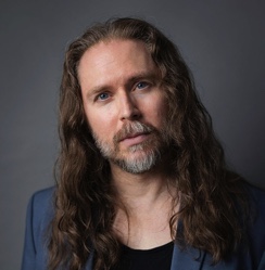 Middle-aged man with long wavy hair, beard, and blue eyes wearing a blue blazer over a black shirt.
