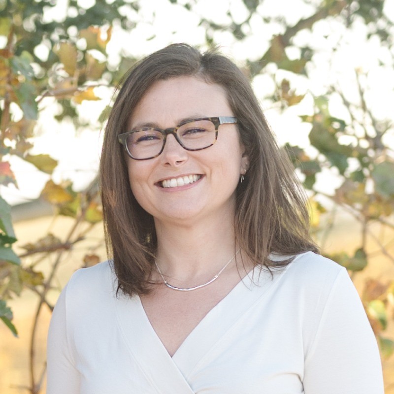 Smiling woman with glasses and brown hair wearing a white top standing outdoors with trees behind her.