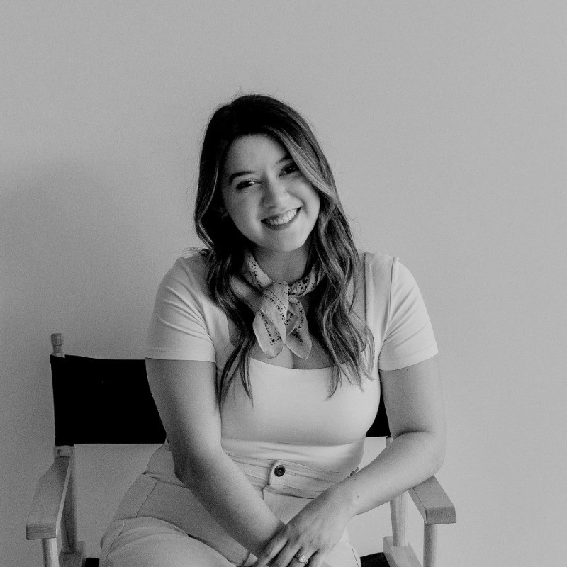 Smiling woman with long hair seated on a director's chair against a plain background.