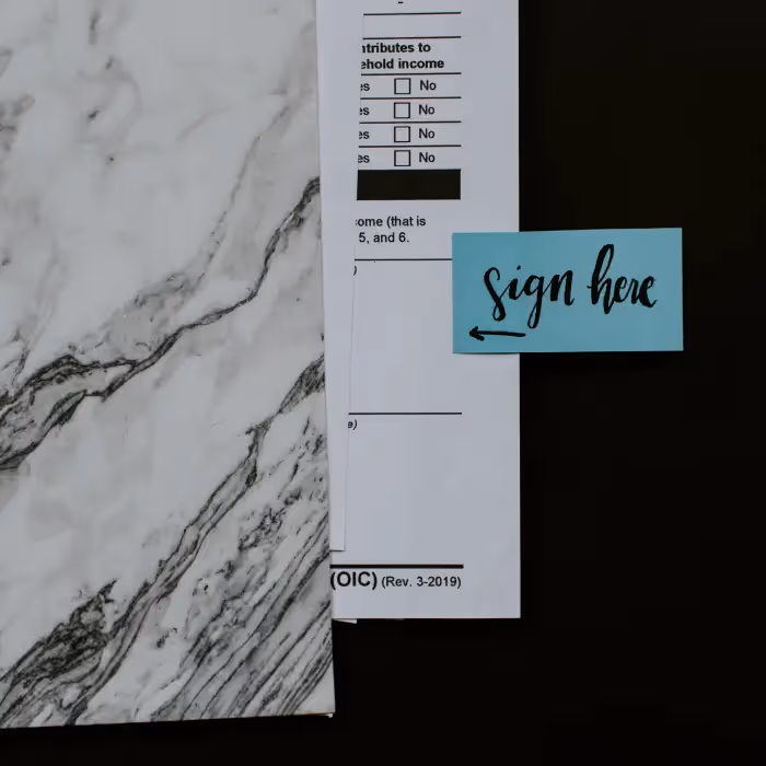 Document corner with a blue sticky note labeled "sign here" next to a marble and black surface.