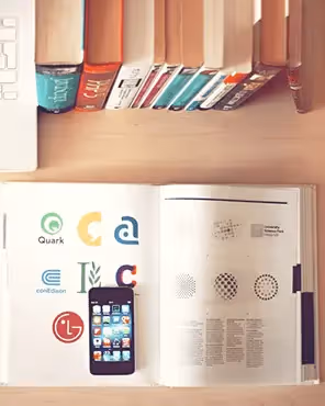 Logo design workspace: Overhead view of wooden desk: white laptop with iPod mini ad, stack of books, sketchpad & pencils, logo design book and smartphone.