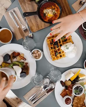 Overhead shot of a round table set for brunch: three plates of waffles with berries and cream, eggs dishes, toast, cups of coffee, glasses of water, and cutlery.