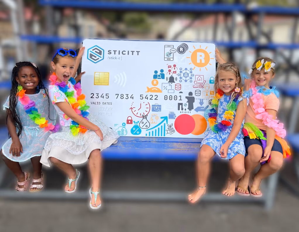 Four young school girls wearing colorful flower leis sit on a blue bench, smiling as they hold up a large, stylised Sticitt Mastercard.