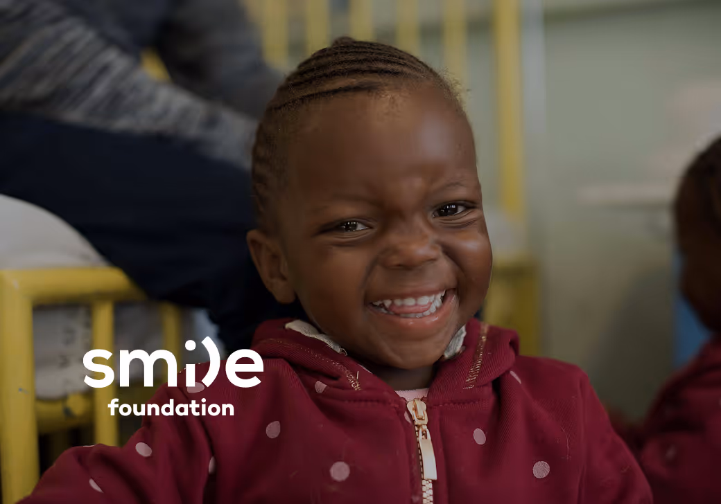 A close-up, joyful portrait of a young child with a wide smile, wearing a maroon jacket. The Smile Foundation logo is overlaid in white in the bottom left corner.