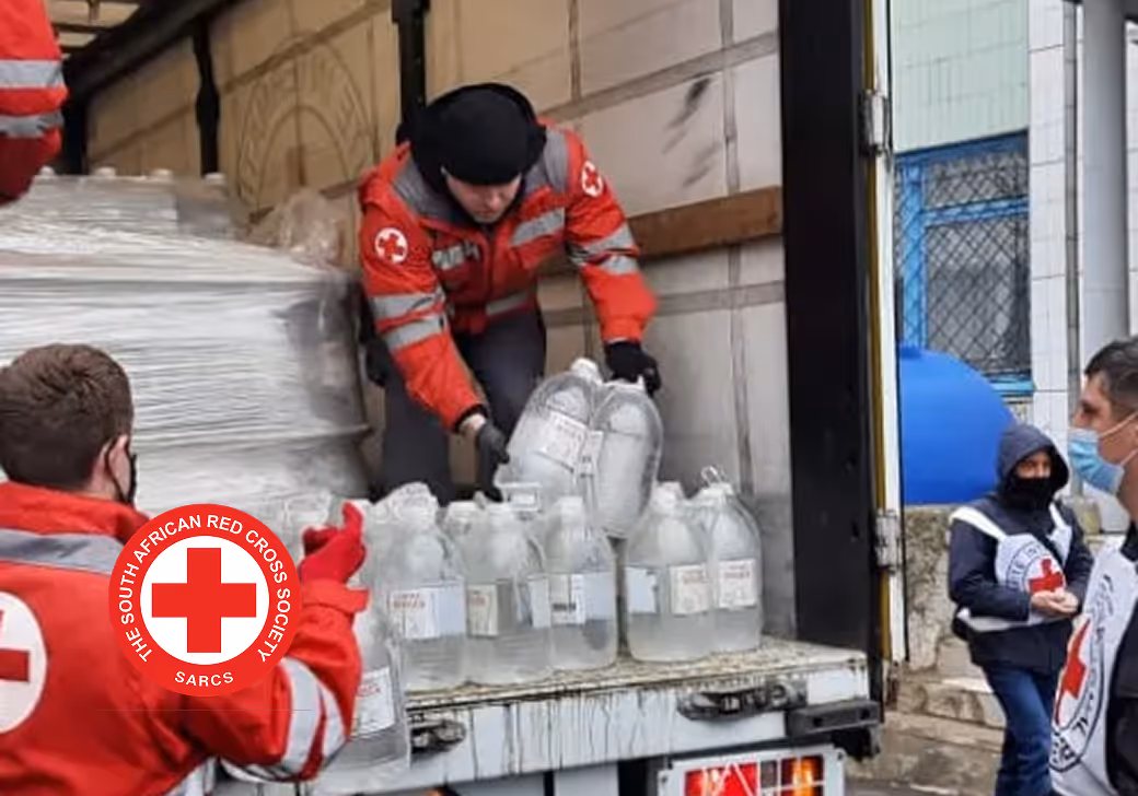Red Cross volunteers in red uniforms unloading large containers of water from the back of a truck. The South African Red Cross Society logo is visible on the side of the vehicle.