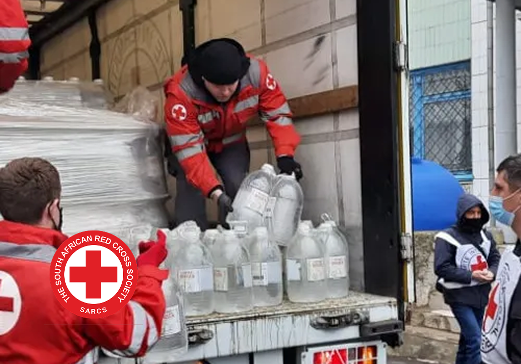 Red Cross volunteers in red uniforms unloading large containers of water from the back of a truck. The South African Red Cross Society logo is visible on the side of the vehicle.
