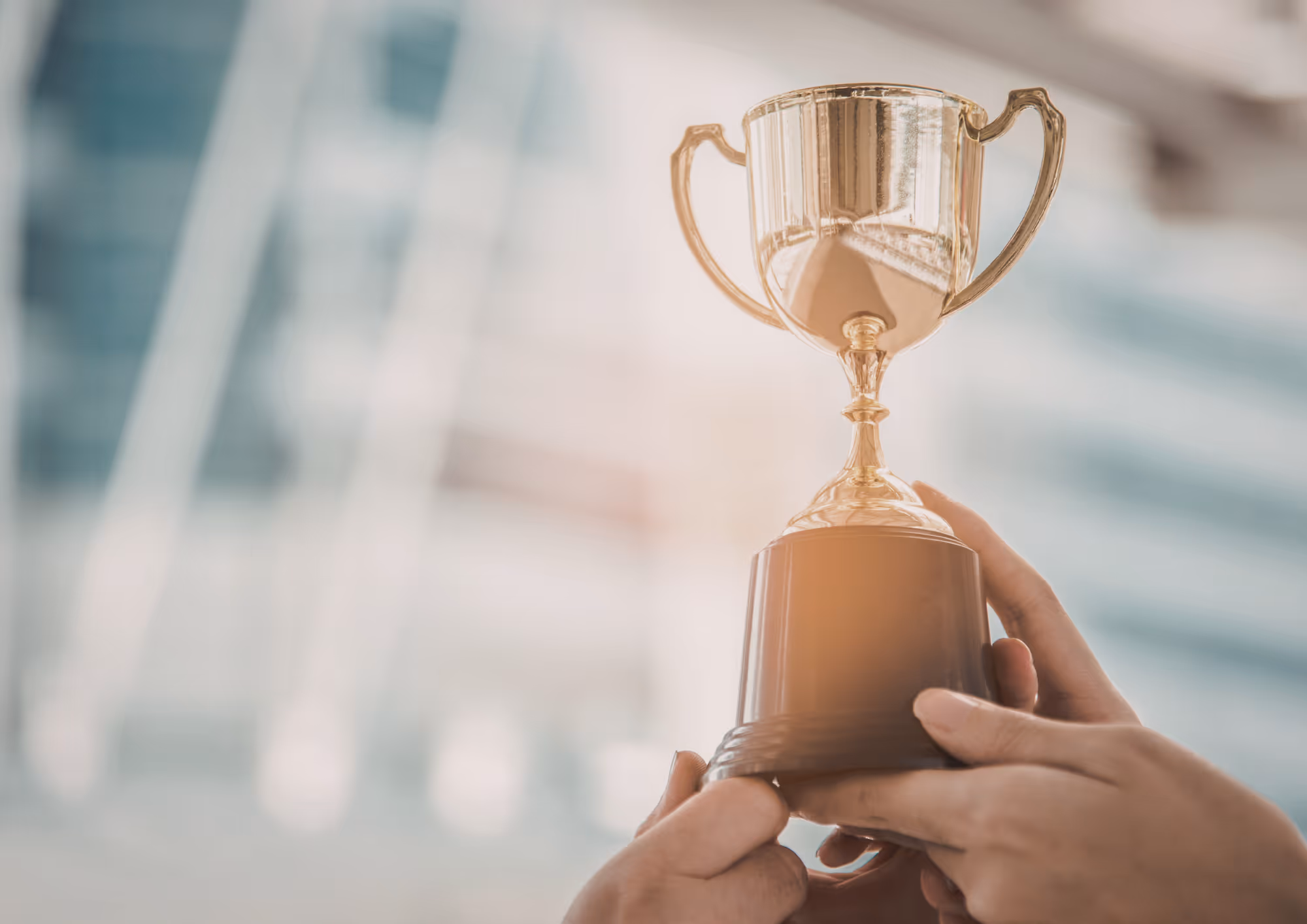 A close-up shot of a person's hands holding up a gold trophy against a blurred urban background with soft sunlight hitting the cup.