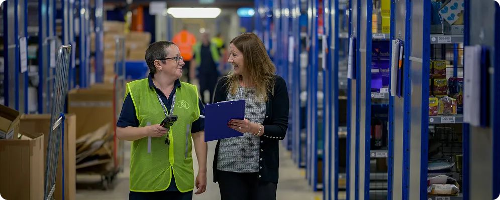 Two women smiling and talking while walking through a warehouse aisle with shelves stocked with boxes.