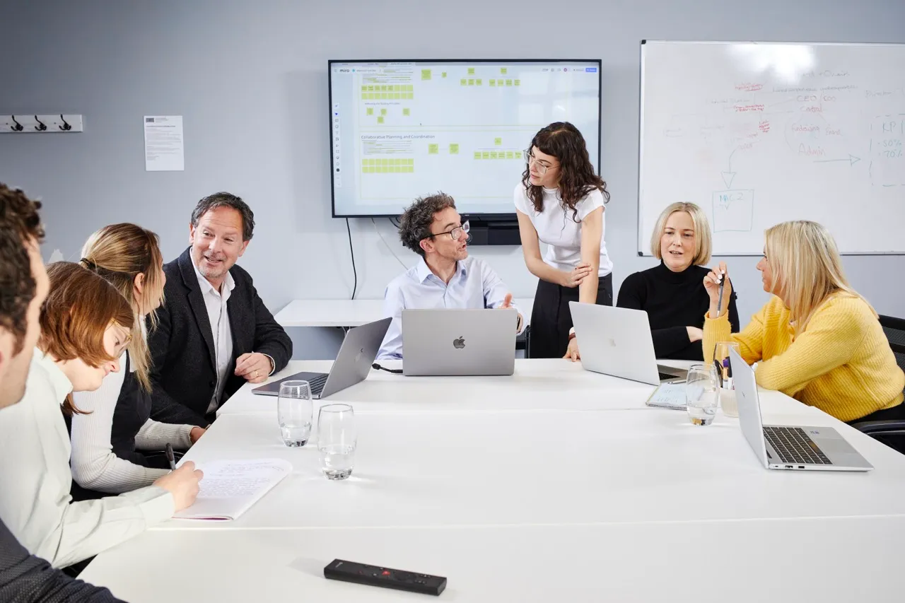 Group of six people in a meeting room discussing around a white table with laptops, a screen showing sticky notes, and a whiteboard in the background.