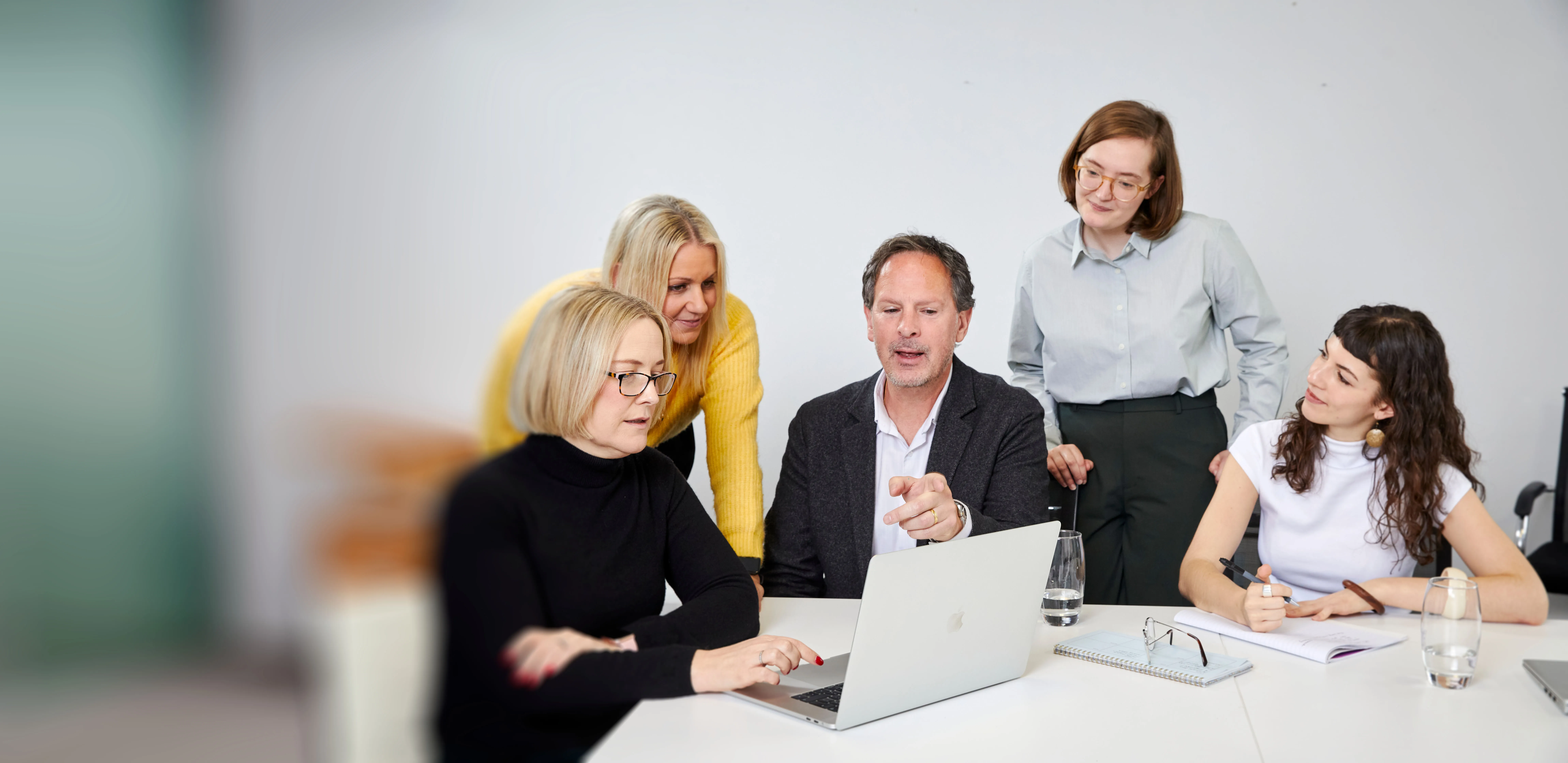 Five colleagues around a table engaged in discussion while viewing a laptop screen in an office setting.