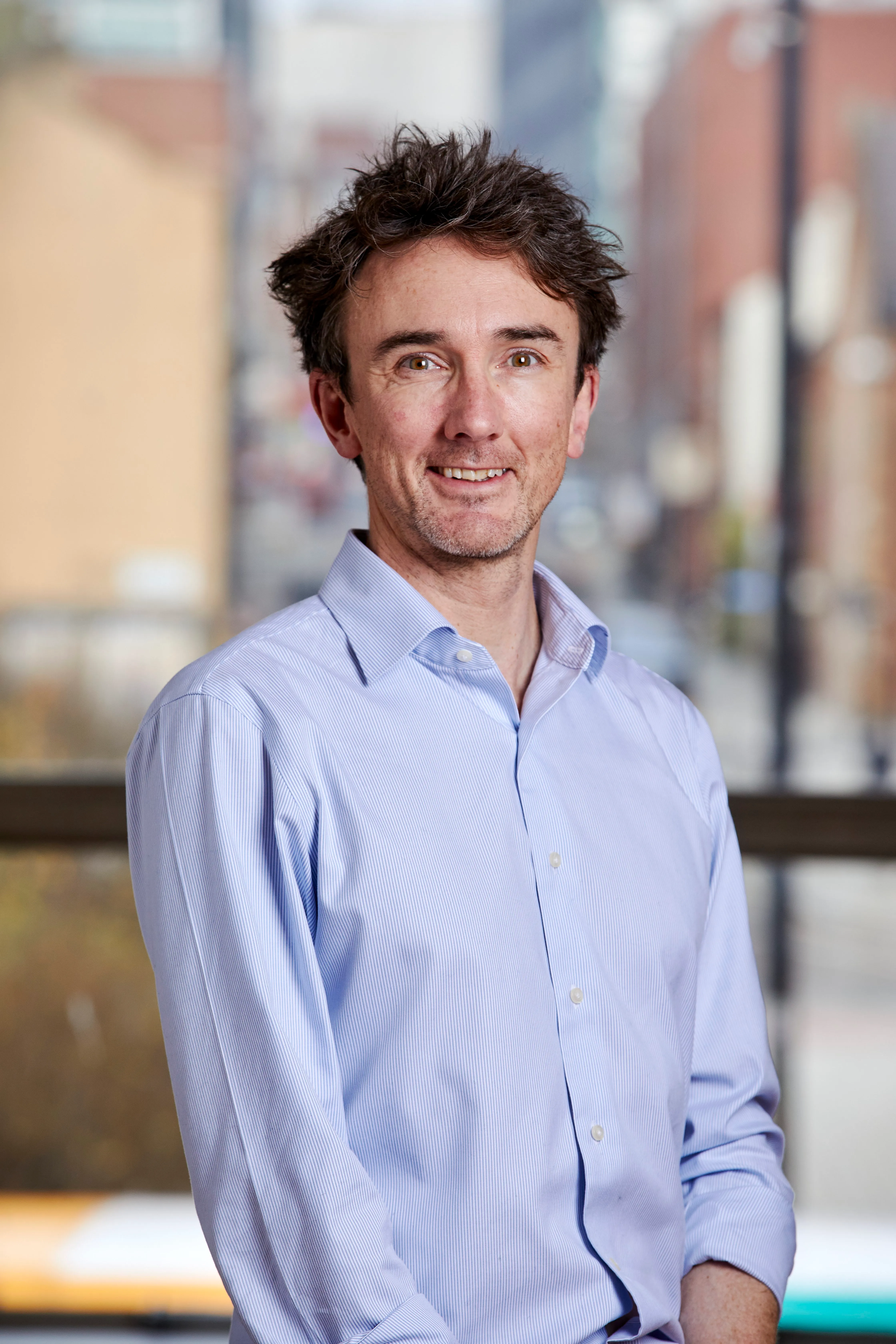 Smiling man with dark curly hair wearing a light blue button-up shirt standing indoors with a blurred background.