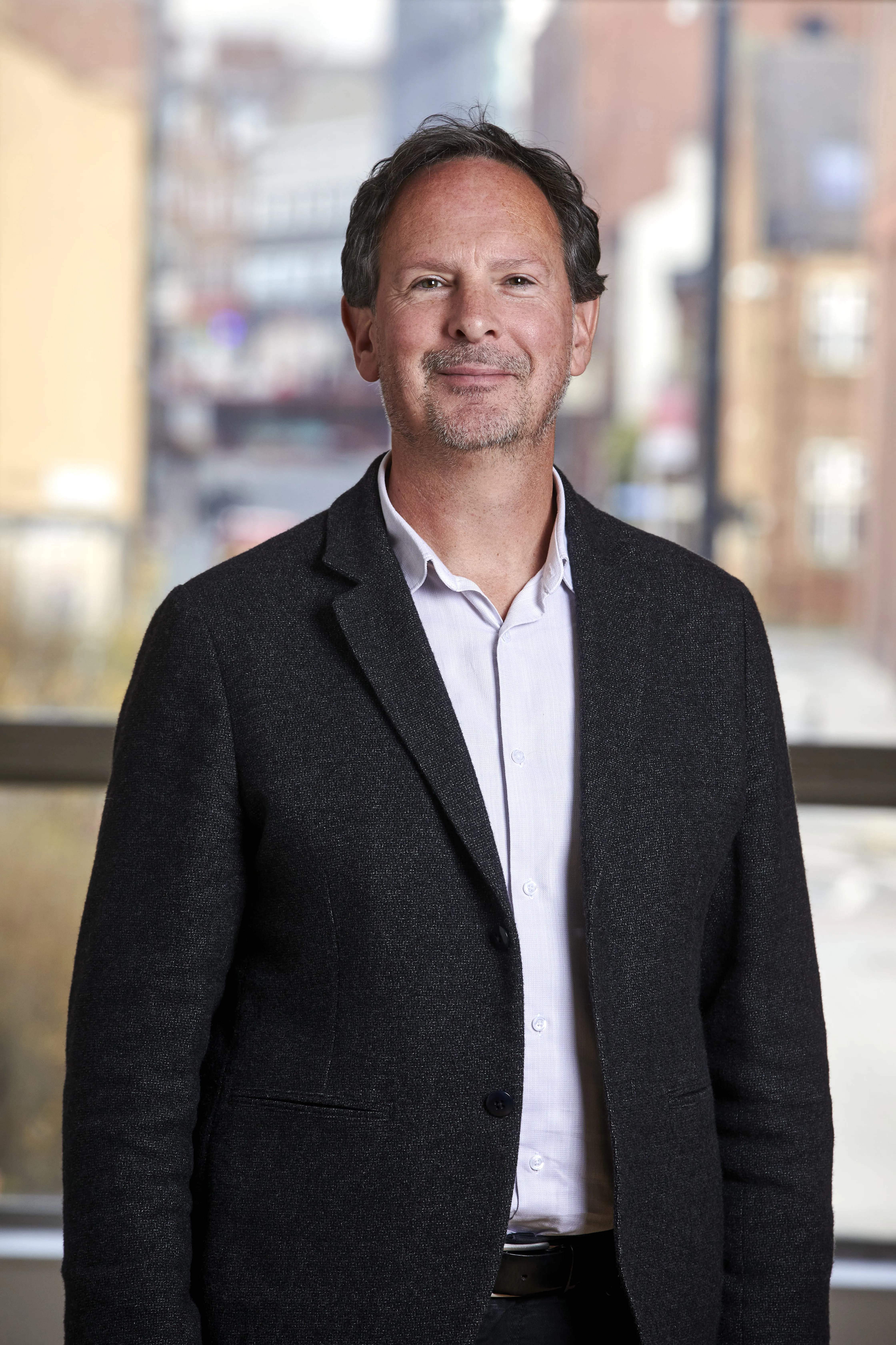 Smiling middle-aged man with short dark hair and stubble, wearing a dark blazer over a white shirt, standing indoors with a blurred cityscape background.
