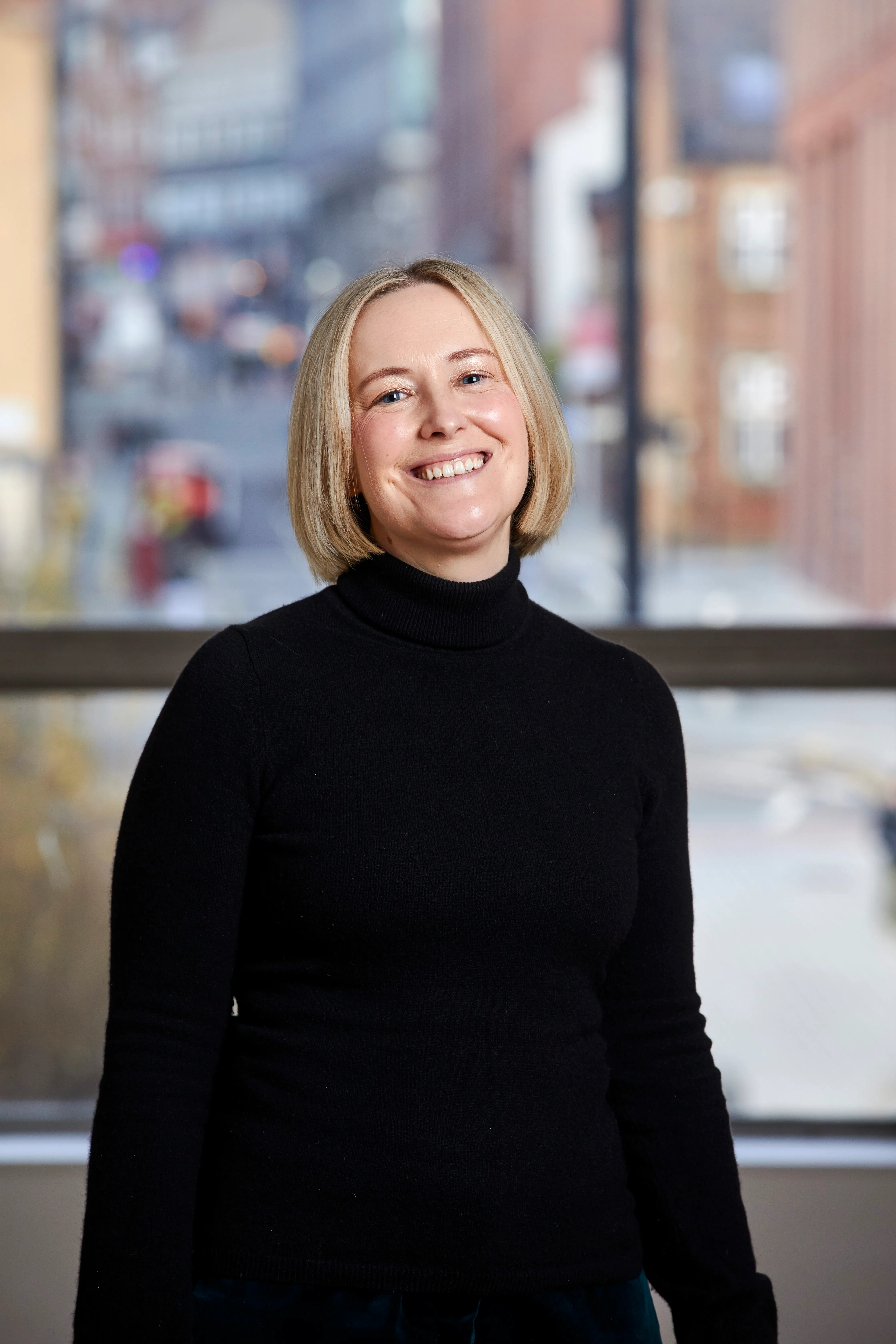 Smiling woman with blonde hair wearing a black turtleneck sweater standing indoors with a blurred city street visible through the window behind her.