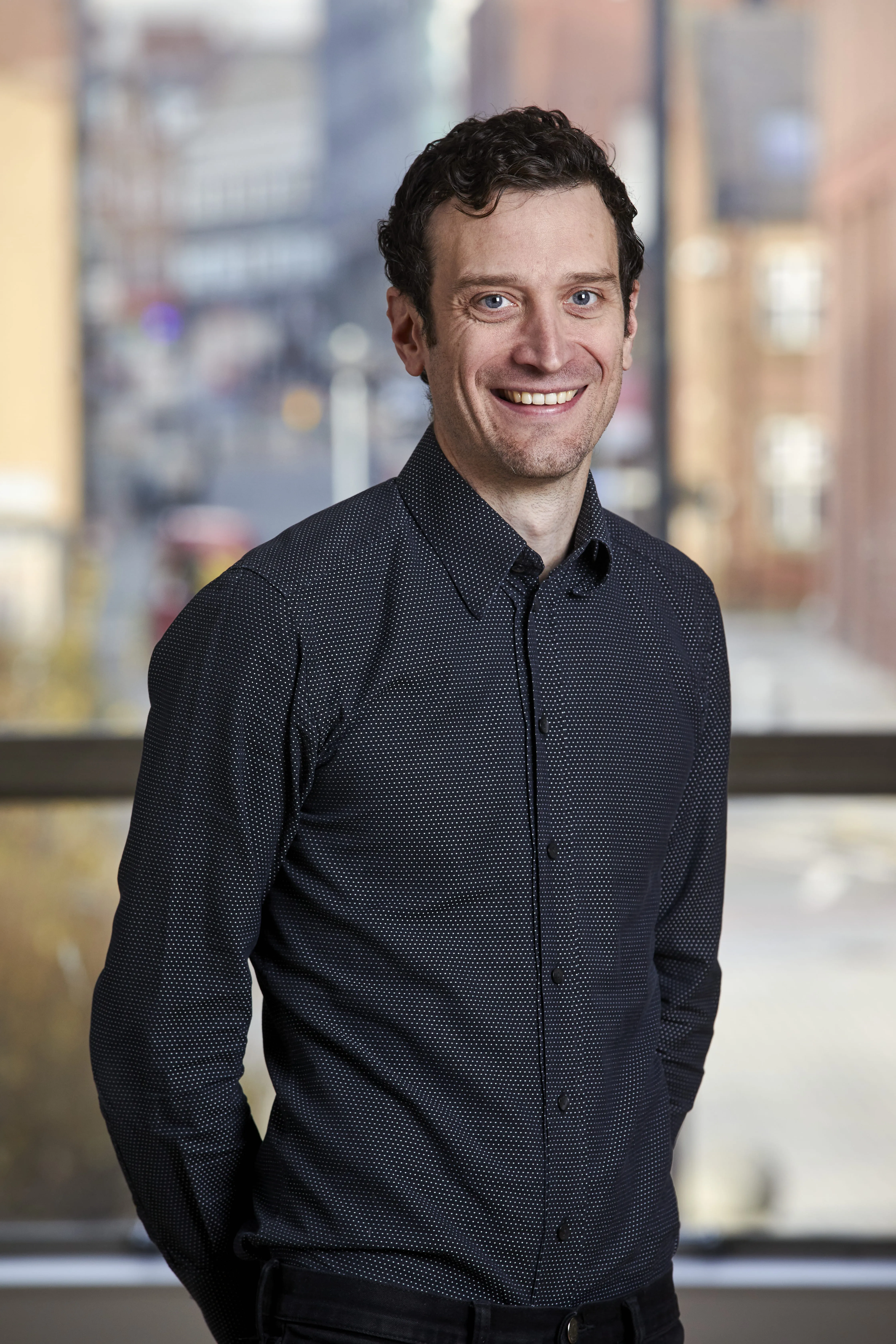Smiling man with curly dark hair wearing a black button-up shirt standing indoors with a city view behind.