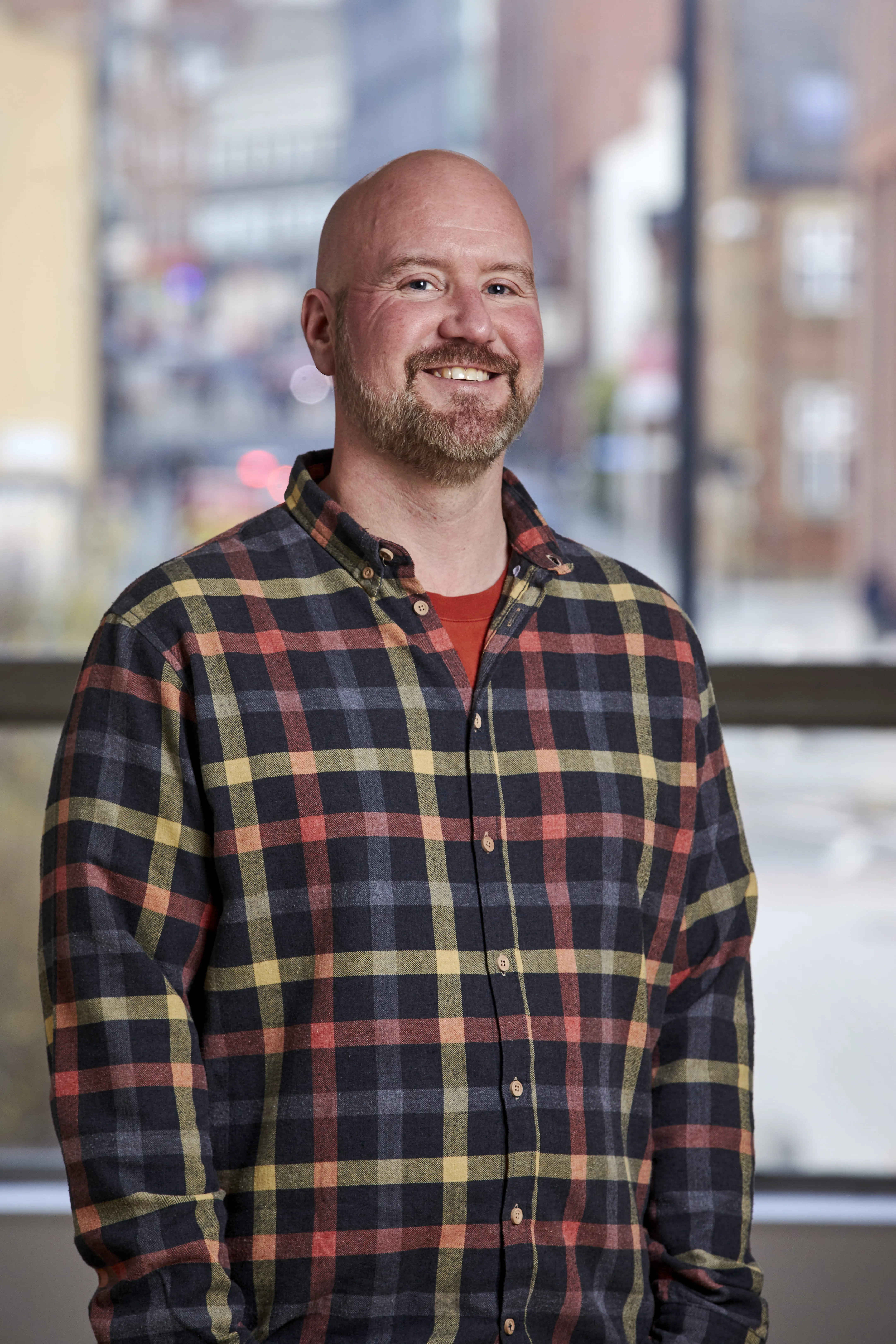 Smiling bald man with a beard wearing a multicolored plaid shirt standing indoors in front of a large window with blurred city buildings in the background.