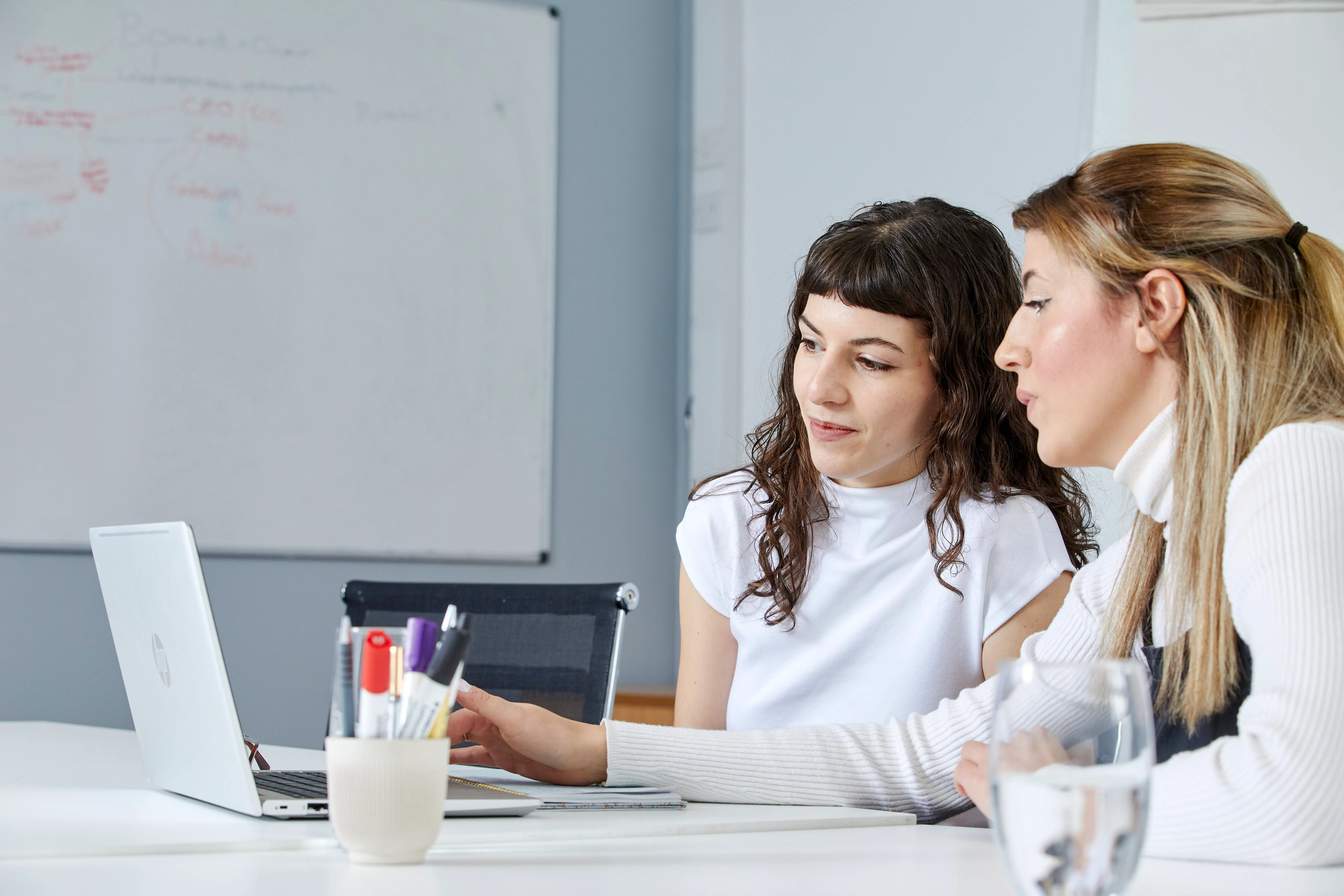 Two women sitting at a table in an office, looking at a laptop screen with a whiteboard in the background.