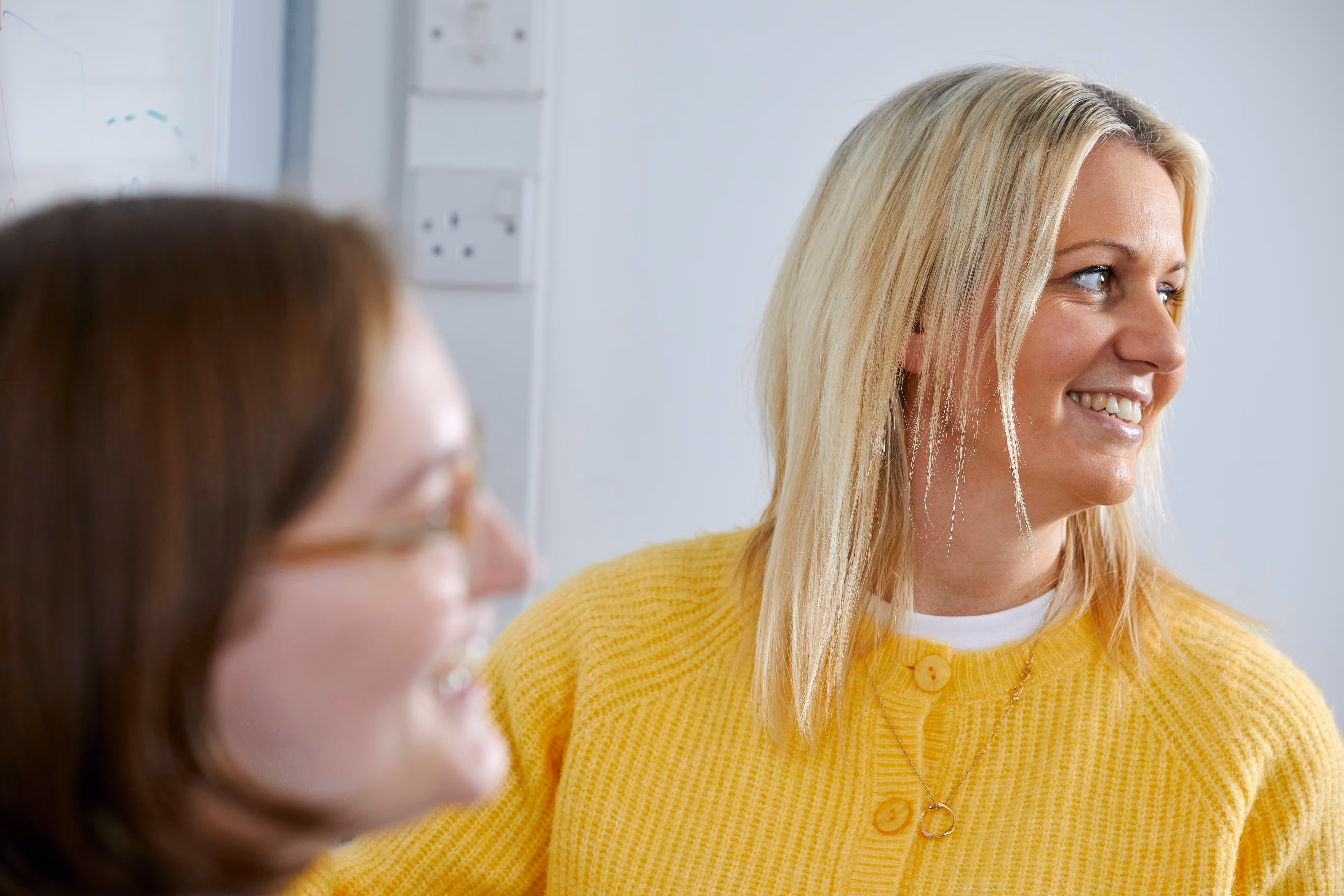 Two women smiling, one with blonde hair wearing a yellow sweater in focus, the other with brown hair and glasses blurred in the foreground.