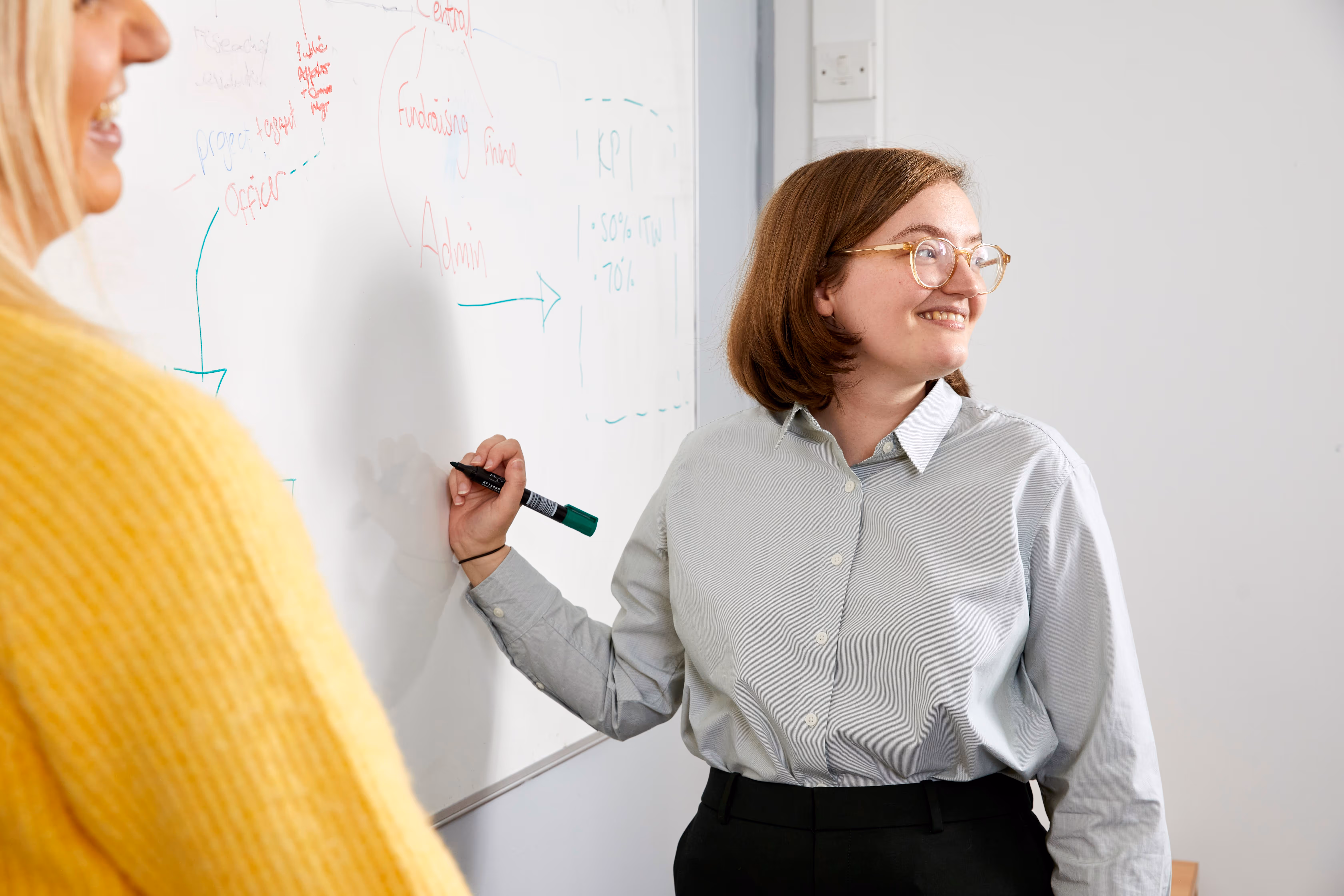 Smiling woman with glasses writing on a whiteboard while another person in a yellow sweater watches.