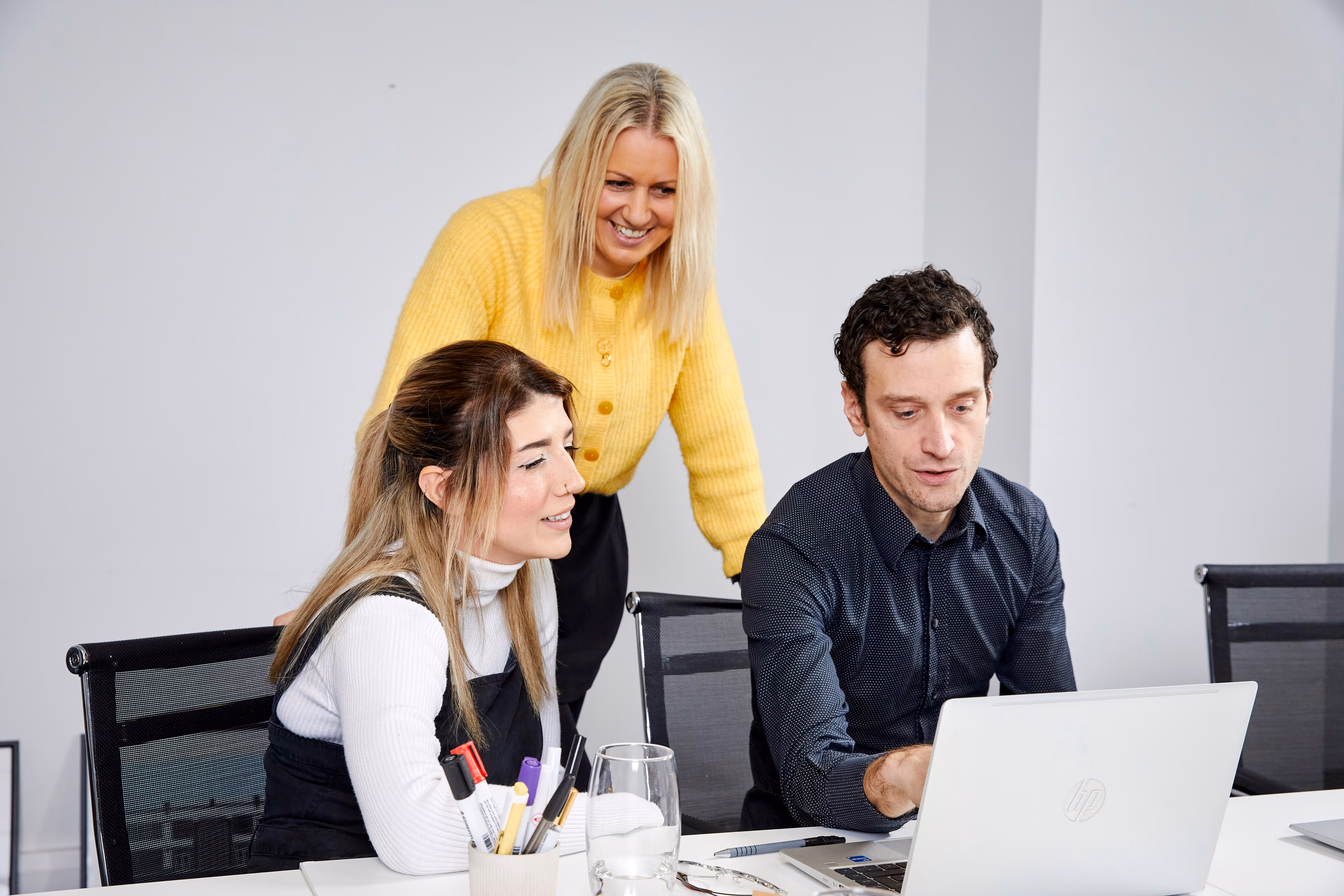 Three colleagues collaborating around a laptop in an office with a white background, one woman standing and smiling, two seated and focused on the screen.