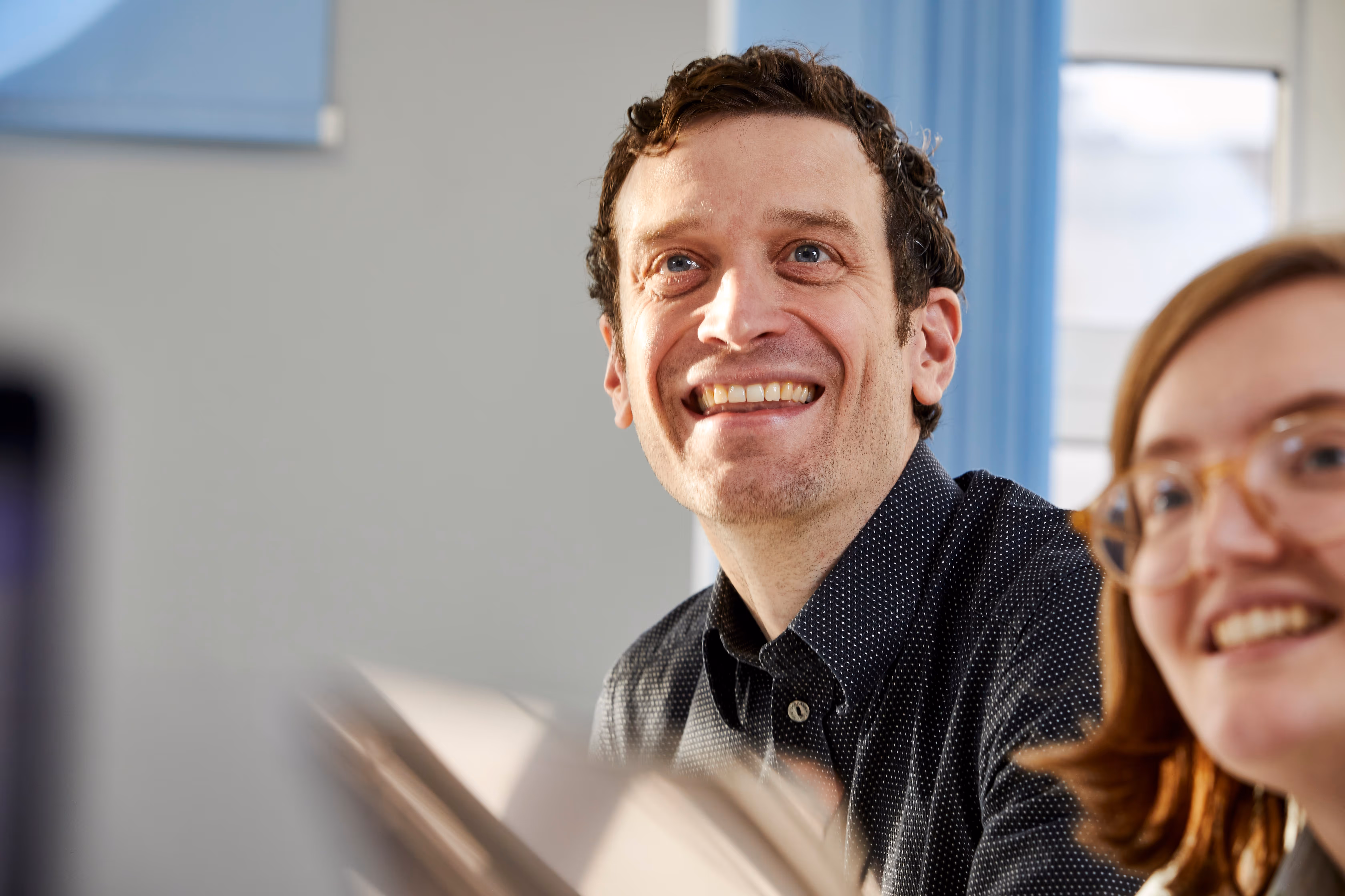 Smiling man with curly hair and a black patterned shirt looking slightly to the right, with a woman wearing glasses partially visible in the foreground.