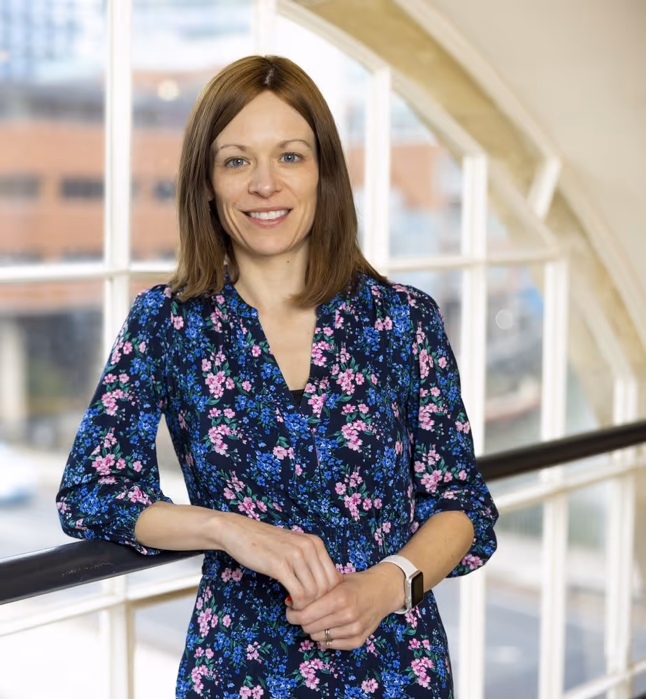 Man with dark curly hair wearing a light blue dress shirt smiling in a bright indoor setting.
