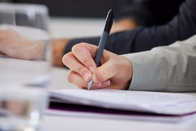 Three colleagues in an office gathered around a laptop, with one man pointing at the screen explaining something.