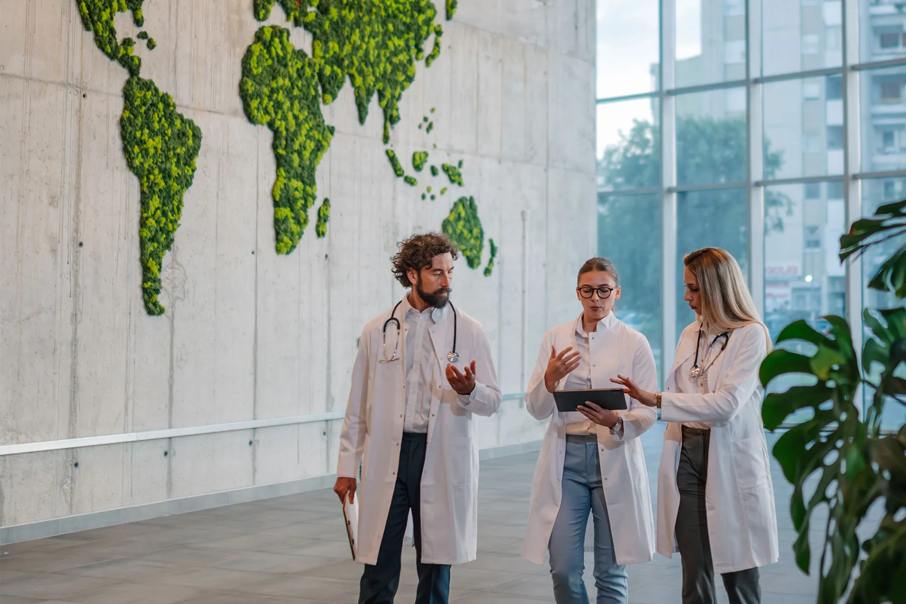 Three doctors in white coats having a discussion in a modern building with a green world map on the wall.