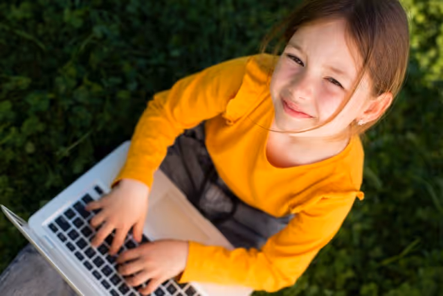 Smiling girl in a yellow shirt using a laptop outdoors on green grass.