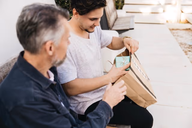 Young man sitting on a bench outdoors, opening a blue gift box while an older man watches.