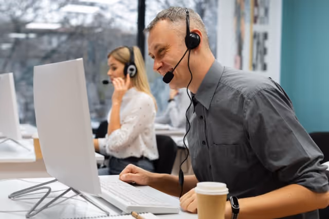 Man and woman wearing headsets working at computers in a modern office with a coffee cup on the desk.