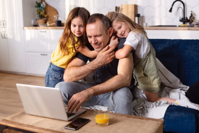 Father working on laptop at home while two young daughters hug him from behind on a couch.