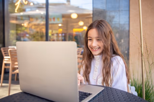 Smiling young girl with long hair sitting at an outdoor table using a laptop.