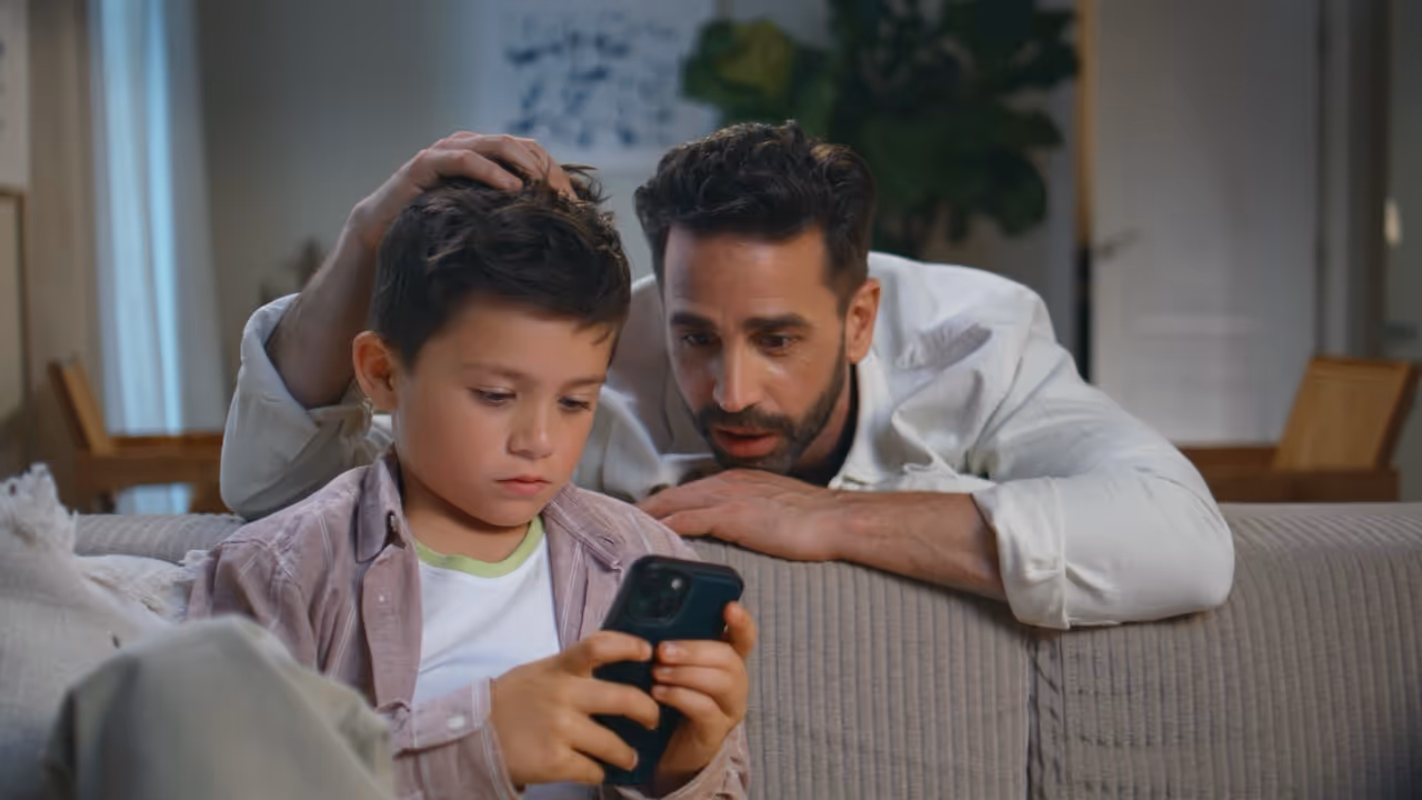 Serious young boy playing on a smartphone while a bearded man watches over him on a couch in a cozy living room.