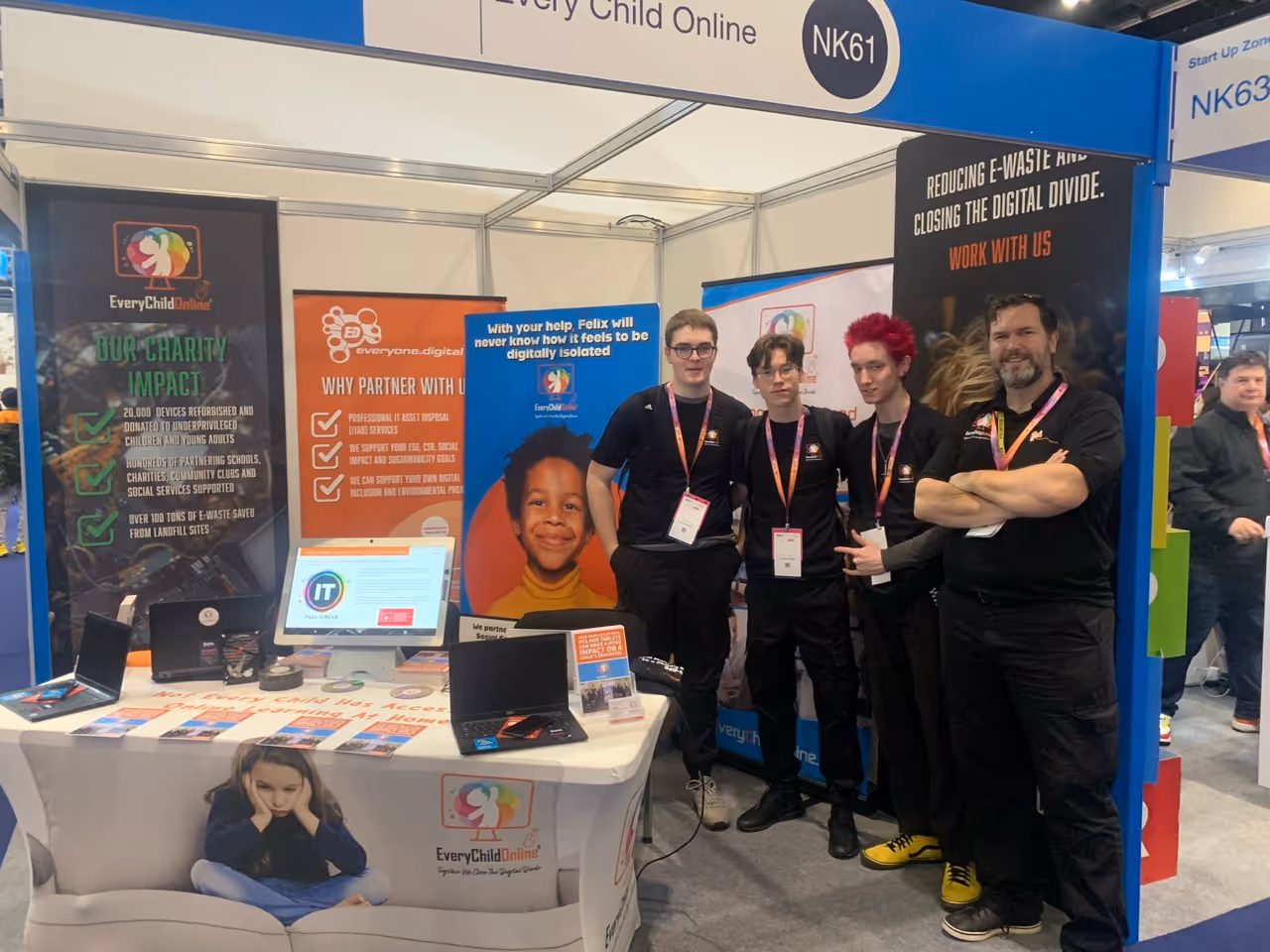 Four people wearing black shirts with lanyards standing at a booth with banners about digital inclusion and charity impact.
