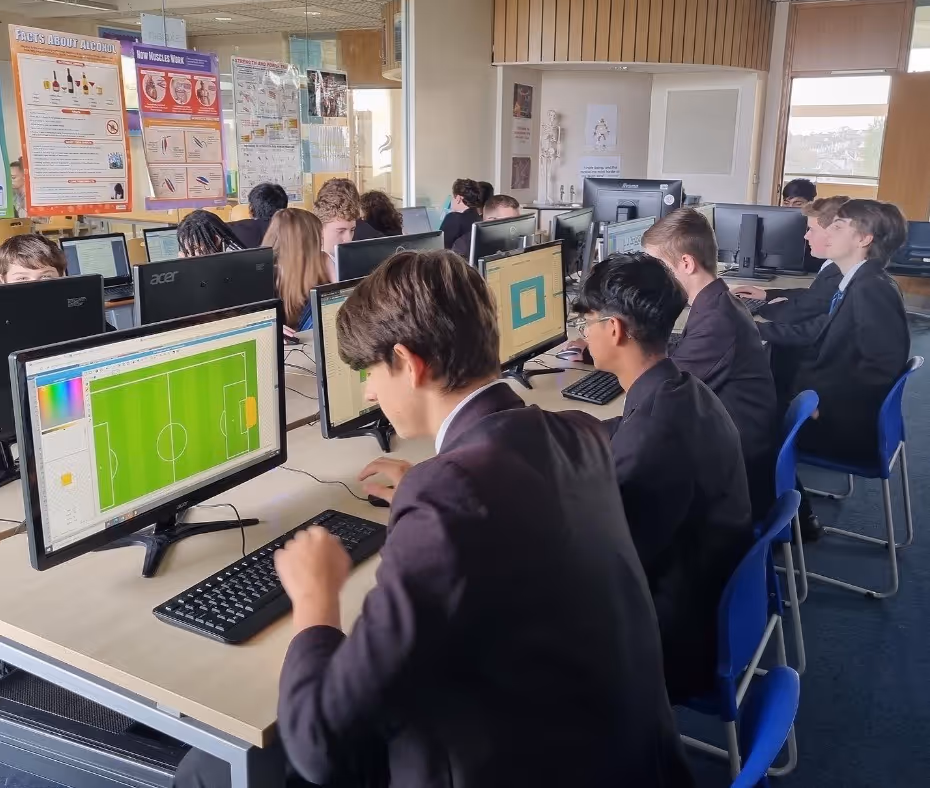 Students in school uniforms working on desktop computers in a classroom with educational posters on the walls.