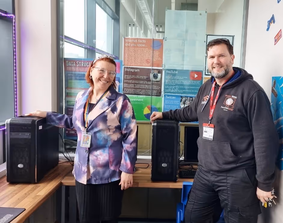 Two people smiling and standing beside desktop computer towers on a wooden table in a room with technology posters on the glass wall behind.