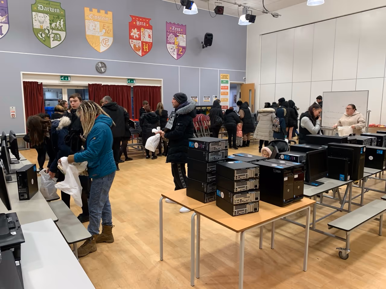 People in winter clothing inside a hall picking up desktop computers stacked on tables.
