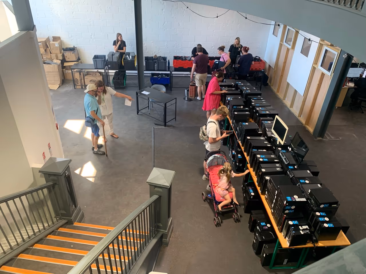 People in a large indoor space examining rows of black desktop computer towers and monitors on tables.