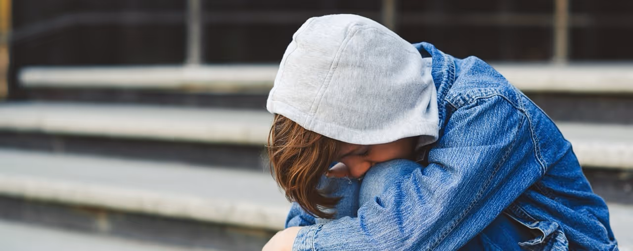 Person wearing a gray hoodie and blue denim jacket sitting on steps with head resting on bent arms.