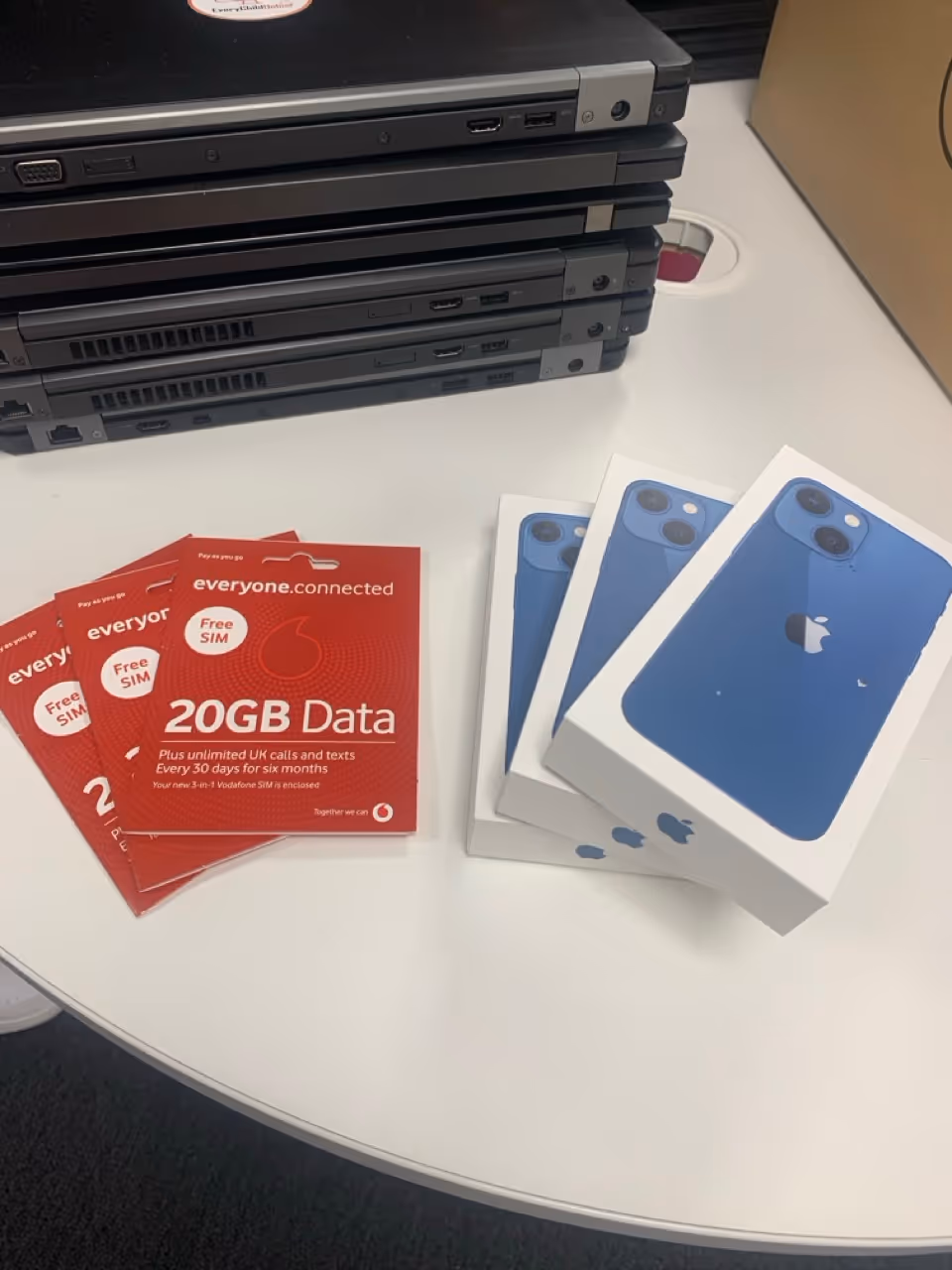 Stack of laptops on a table next to three blue iPhone boxes and three red Vodafone 20GB Free SIM card packages.