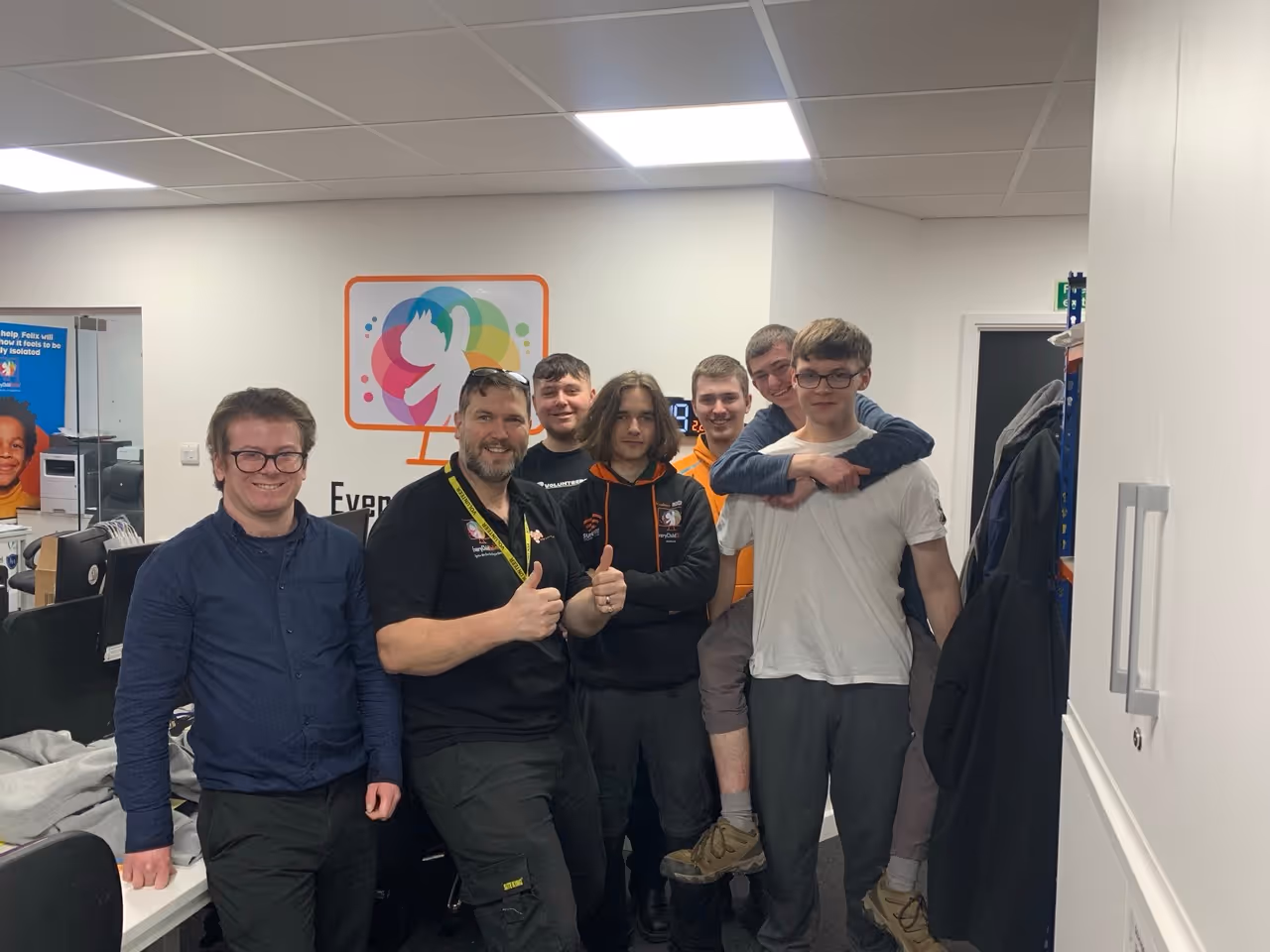 Group of seven men smiling and posing inside an office with a colorful logo on the wall behind them.