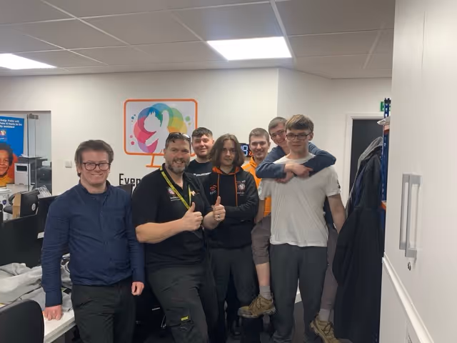 Group of seven men standing and posing in an office, some smiling and one giving thumbs up.