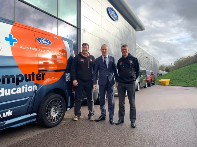 Three men standing on a driveway next to a Ford branded van and a Ford building, with two men in black hoodies and one man in a suit.