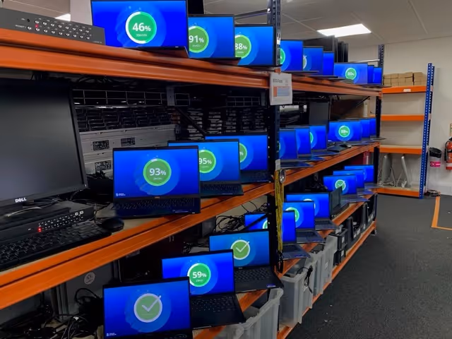 Shelf filled with multiple laptops displaying blue screens showing percentage progress circles in a storage or testing room.