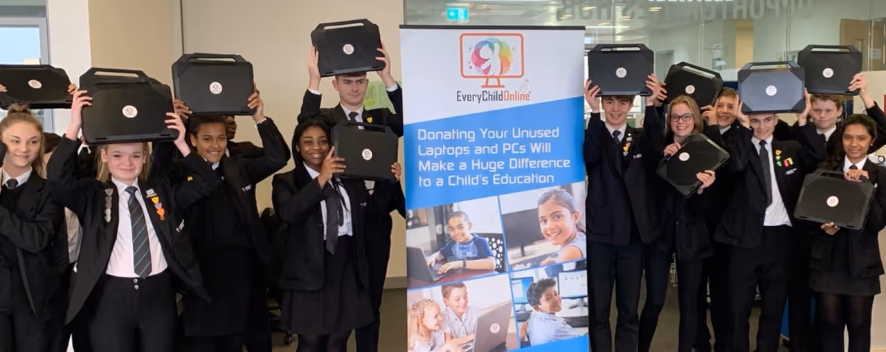 Group of smiling students in school uniforms holding laptops above their heads standing next to an EveryChildOnline banner promoting laptop donations for children's education.