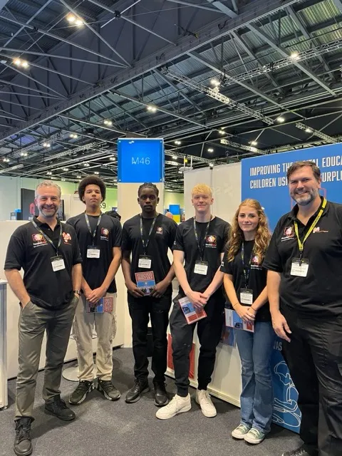 Group of five people standing indoors at a trade show booth, wearing black shirts with name badges.